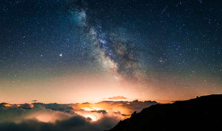 Starry sky above a sea of clouds with the lights of the cities and villages below producing orange...