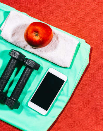 Attributes of a healthy lifestyle: apple, sports equipment, hand weights, sports shirt on a red mat for sport in background. Flat view with copy space.の写真素材