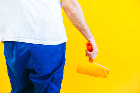Anonymous painter man at work painting a orange wall with a roller, rear view. Offering professional painting services.の写真素材