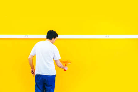 Painter man at work painting a orange wall with a roller, rear view. Offering professional painting services. Isolated on big empty space for copy space.の写真素材