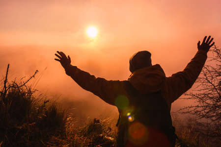 Young adventurous guy raising his hands to enjoy freedom. Traveler backpacker man sitting observing the sun on a beautiful sunset or sunrise. Concept of traveling and adventure.の写真素材