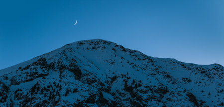 Snowy mountain peaks with the moon at blue hour. Nature background landscape. Moon over the snowy mountains with blue and clear sky on early morning in the mountains. Panoramic scenery.の写真素材