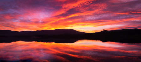 Sunrise or sunset with orange, purple and red clouds reflected in the lake water. Natural mirror at golden hour. Spectacular and beautiful scenery. Estany de Banyoles, Girona, Catalonia, Spain.の写真素材