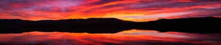 Panoramic sunrise or sunset with orange, purple and red clouds reflected in the lake water. Natural mirror at golden hour panorama. Spectacular and beautiful scenery.の写真素材
