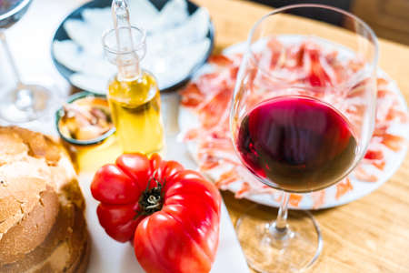 Typical Catalan or Spanish food. Bread with tomato, Iberian ham, glass of red wine, goat cheese, oil and garlic. Food products for dinner or tapas on a wooden background.の写真素材