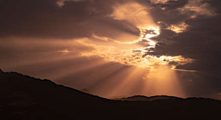 Rays of light filtering through the clouds in a beautiful sunset or sunrise behind mountain silhouette. Picturesque view of sky with sun rays, beautiful natural orange and warm light.の写真素材