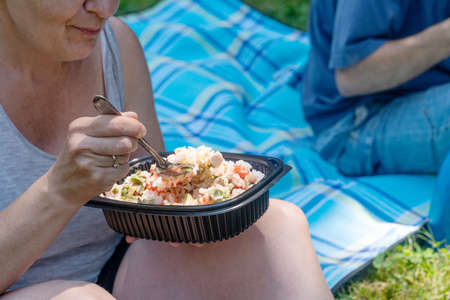 Anonymous woman eating homemade food prepared in a reusable plastic lunch box in the field. Summer vacation, eating outdoors.の写真素材