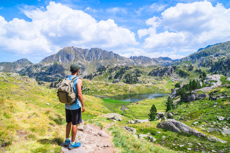 Young man with a bag standing on an alpine mountain trail with a lake.の写真素材