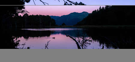 Picturesque sunset or sunrise of beautiful landscape with illuminated peaks, silhouette of some ducks and trees. Fantastic evening panorama of alpine mountain lake. Beauty of nature concept.の写真素材