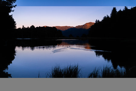 Picturesque sunset or sunrise of beautiful landscape with illuminated peaks, silhouette of some ducks and trees. Fantastic evening panorama of alpine mountain lake. Beauty of nature concept.の写真素材
