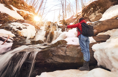Beautiful girl in red jacket with black backpack stands arms up in front of the winter waterfall and orange sunの写真素材
