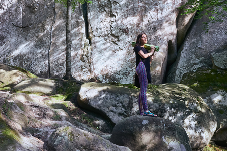 Young attractive girl standing on the big rock in mountain. Preparing for practicing yoga on a yoga mat.の写真素材