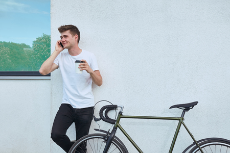 Young hipster style man posing with bicycle on the street sport style picture handsome guy white wallの写真素材