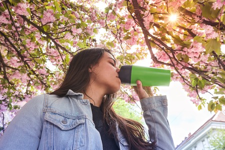 Young girl drinking and enjoying in sunshine standing outdoor in sun light on the background of blooming sakuraの写真素材