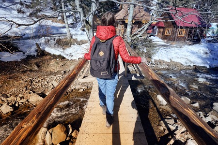 A happy girl, with a red jacket and a black backpack, crossing a wooden bridge, during a winter hiking trip, with snow covered trees around her.の写真素材