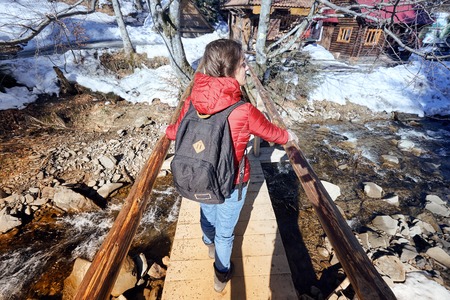 A happy girl, with a red jacket and a black backpack, crossing a wooden bridge, during a winter hiking trip, with snow covered trees around her.の写真素材