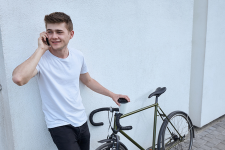 Young hipster style man posing with bicycle on the street sport style picture handsome guy white wallの写真素材