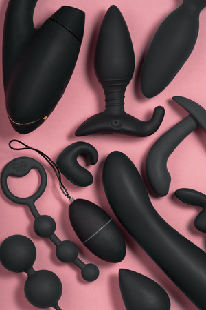 Erotic pleasure toys on a pink background. Sex gadgets and masturbation devices. Black dildo surrounded by black rubber and silicone sex butt plugs, vibrators, vaginal balls. Top view.の写真素材