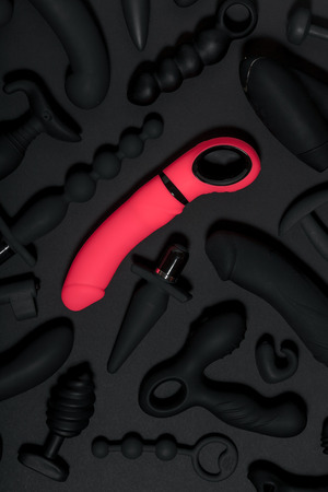 Erotic pleasure toys on a black background. Sex gadgets and masturbation devices. Pink dildo surrounded by black rubber sex toys: anal butt plugs, vibrators, vaginal balls. Top view.の写真素材