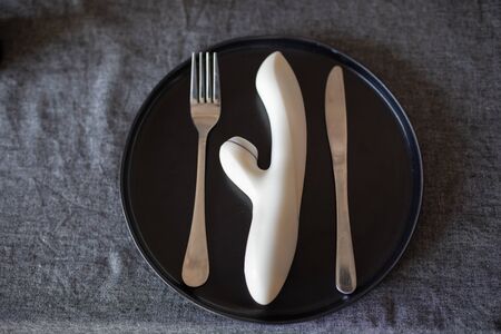 A white vibrator lies on a black plate between the fork and the knife. Dildo for women, stimulating the vagina and clitoris. Image for sex shop. Sex toys for women. Rabbit dildos with vibration.の写真素材