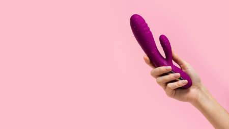 The girl's hand holds a purple vibrator covered with drops on a pink background. Vibrator for female masturbation. Dildo for vaginal and clitoris stimulation. Image for a sex shop. Sex toys for adultの写真素材