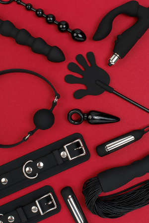 BDSM Erotic pleasure toys on a red background. Sex gadgets and masturbation devices. Black dildo surrounded by black rubber and silicone sex toys: anal butt plugs, vibrators, gag, cuffs and whipの写真素材