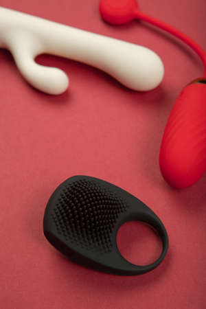 Black-silicone erection ring lies against the background of a vibrator and vaginal balls on burgundy paper. Erotic toy for fun. Sex gadget and masturbation device. View from the side. Place for text. Sex shop concept.の写真素材