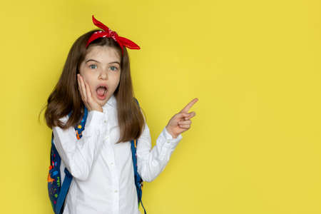little beautiful school girl in a school uniform with a backpack surprised, opened her mouth. Advertising concept back to school, September 1. Portrait on a yellow backgroundの写真素材