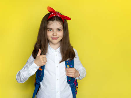 Little beautiful school girl in a school uniform with a backpack raised a finger up. Advertising concept back to school, September 1st, school sale. Portrait on a yellow backgroundの写真素材