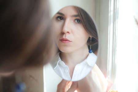 young beautiful girl with brown eyes and a blue earring looks in the mirror. Portrait reflection in...