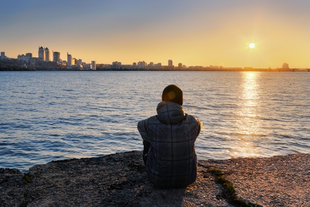 The guy meets the sunset siting on the river bank and looking away into the cityの写真素材