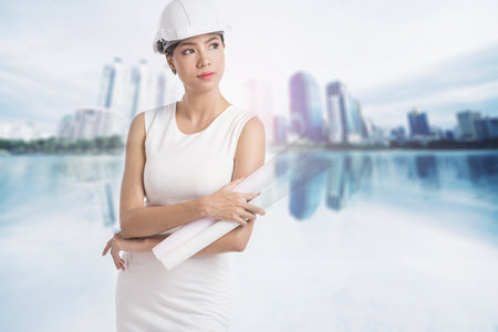 Female engineer crossing her arms and holding paper rolls on blurry buildings backgroundの写真素材