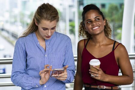 White woman is using mobile phone and talking to smiling black woman that she is holding a cup of...