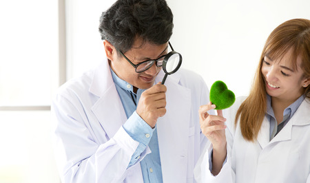 Male scientist holding a magnifying glass and green heart in her hand on lab a background. Beautiful smiling female doctor or Scientist holding organic heart-shaped. science room.の写真素材