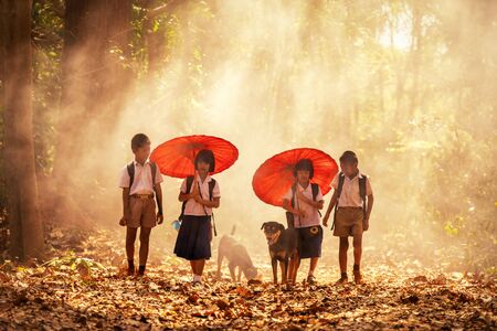 Community life. The boy and girl holding a red umbrella with dog is walking in the forest to go to school. Group of school boys and girls with uniforms in forrest. Back to school. Tha Tum District, Surin, Thailand.の写真素材