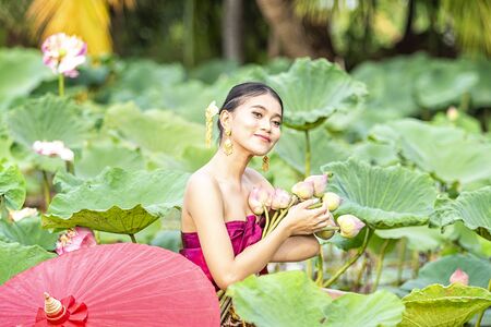 Women wearing Thai dresses are admiring lotus flowers. Asian women sitting on wooden boats to collect lotus. Beautiful girl wearing traditional Thai dress hand holding a flowers.の写真素材