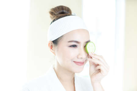 Beauty and spa condo, woman holding cucumber. Young woman with facial mask holding cucumber slices on white background. Copy space.の写真素材