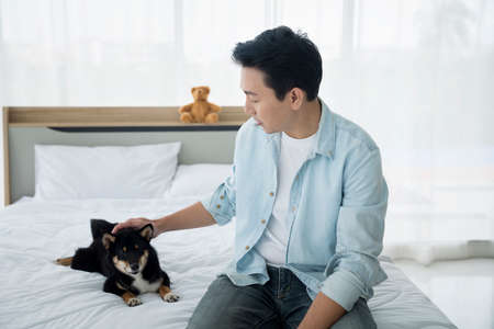 A young man playing with a black Shiba Inu in his bedroom bed.