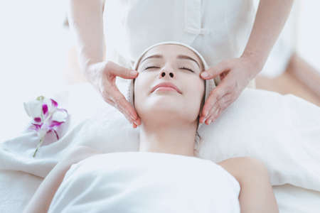 Female massage therapist giving a massage at a spa. Young woman receiving head and facial massage in...