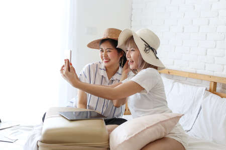 Young Asian couple taking selfie with mobile phone in apartment. Two women are preparing for the journey happily. Young female relaxing In room.の写真素材