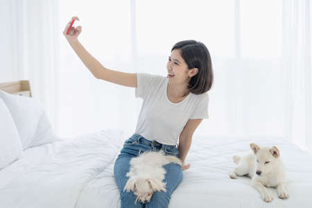 Young Asian women selfie with mobile phone. Young Asian women and girl were in bed with a dog. Girl and white Hokkaido inu dog and Maltese dog.の写真素材