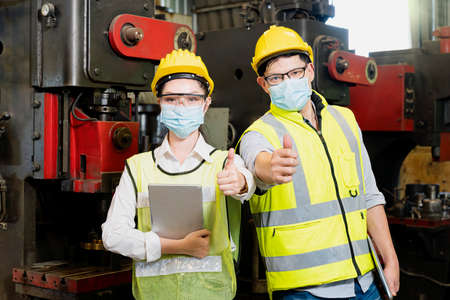 Asian woman and man wearing a mask holding a tablet and show thumb up in an industrial factory. Professional technicians are holding a tablet to control work in industrial plants.の写真素材