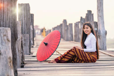 A young Burmese woman wearing traditional clothes with red umbrella at  U Bein teak bridge on Taungthaman lake in Mandalay Division. Myanmar.の写真素材