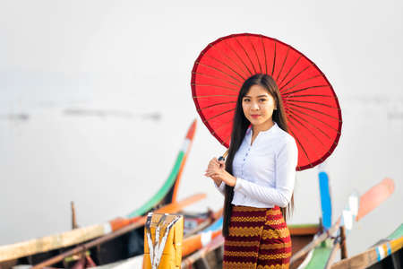 A young Burmese female with a red umbrella on wooden boat at bridge in Asia.U-bein bridge, Mandalay, Myanmar.の写真素材