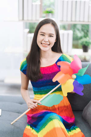 Lgbt Concept. A young woman wearing a rainbow dress is holding a windmill on the sofa in the living room. Lesbian concept.の写真素材