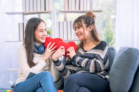 Cute young asian girlfriends in love smiling and holding red hearts on sofa in the living room. Lifestyle of couple loving woman happy in romance love and fun.の写真素材