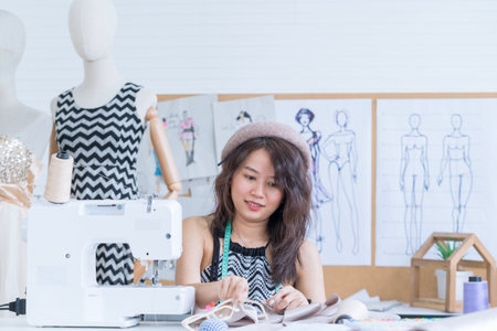 Young fashion designer working on a new dress in her studio. Young Asian worker of a fashionable atelier sitting at the sewing machineの写真素材