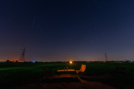 Quadrantid meteor shower. Falling stars over Two chairs and campfire under beautiful night sky full of stars. Rice fields with lamp posts and stars.の写真素材
