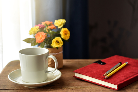 White cup of hot drinks on wooden table by the window. Small vibrant color roses and red cover notebook with pencils on it as background. Hot coffee or tea for a cold morning refreshment.の写真素材