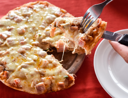Hot cheesy pizza slice being served on wooden plate, red table cloth background.の写真素材